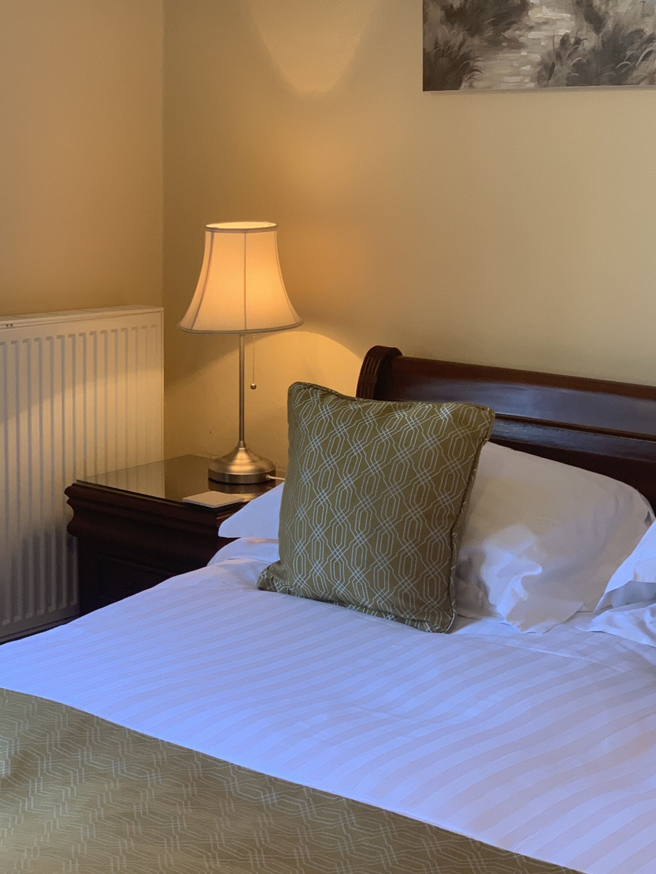 A neatly made bed with a patterned pillow, a bedside lamp, and a radiator.