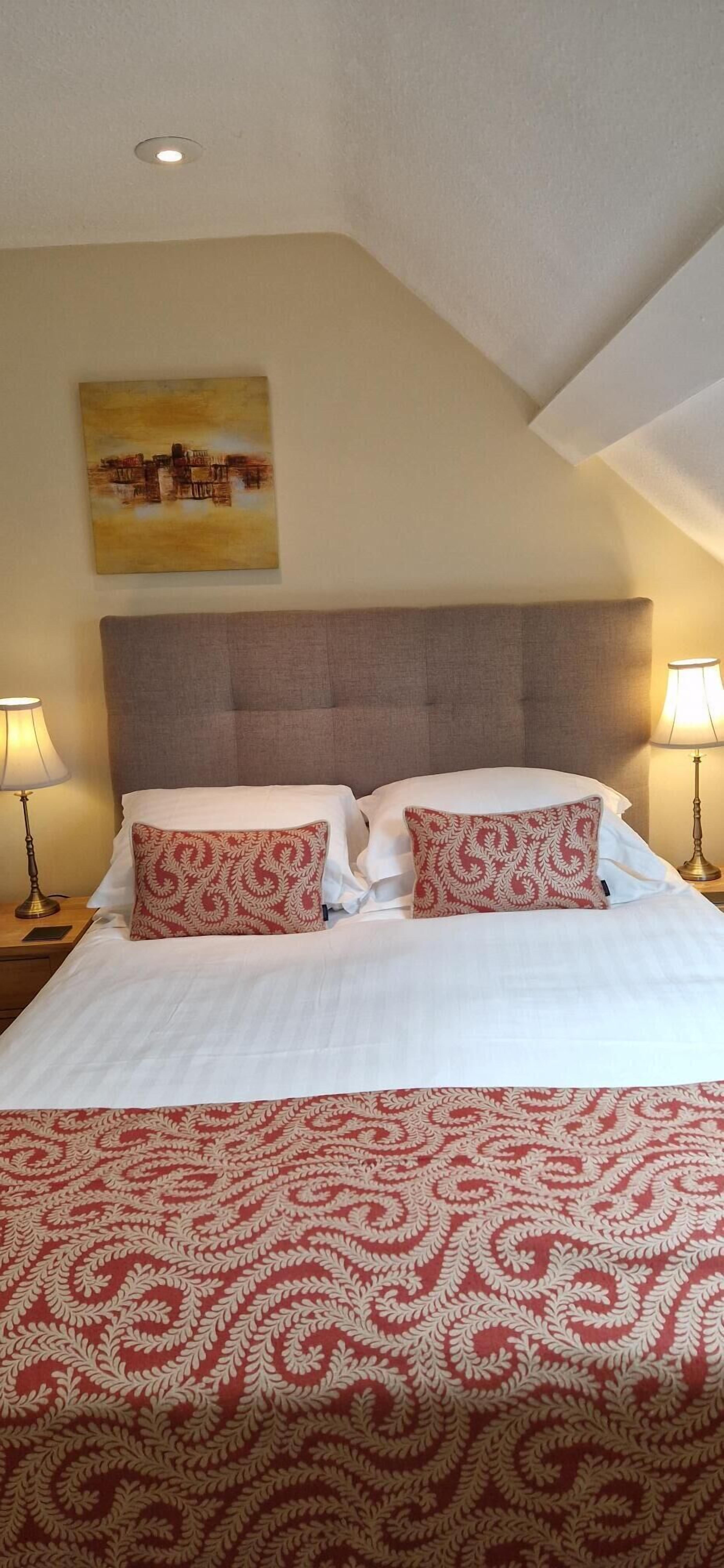 A hotel room with a bed, two bedside lamps, a headboard, and a painting on the wall.