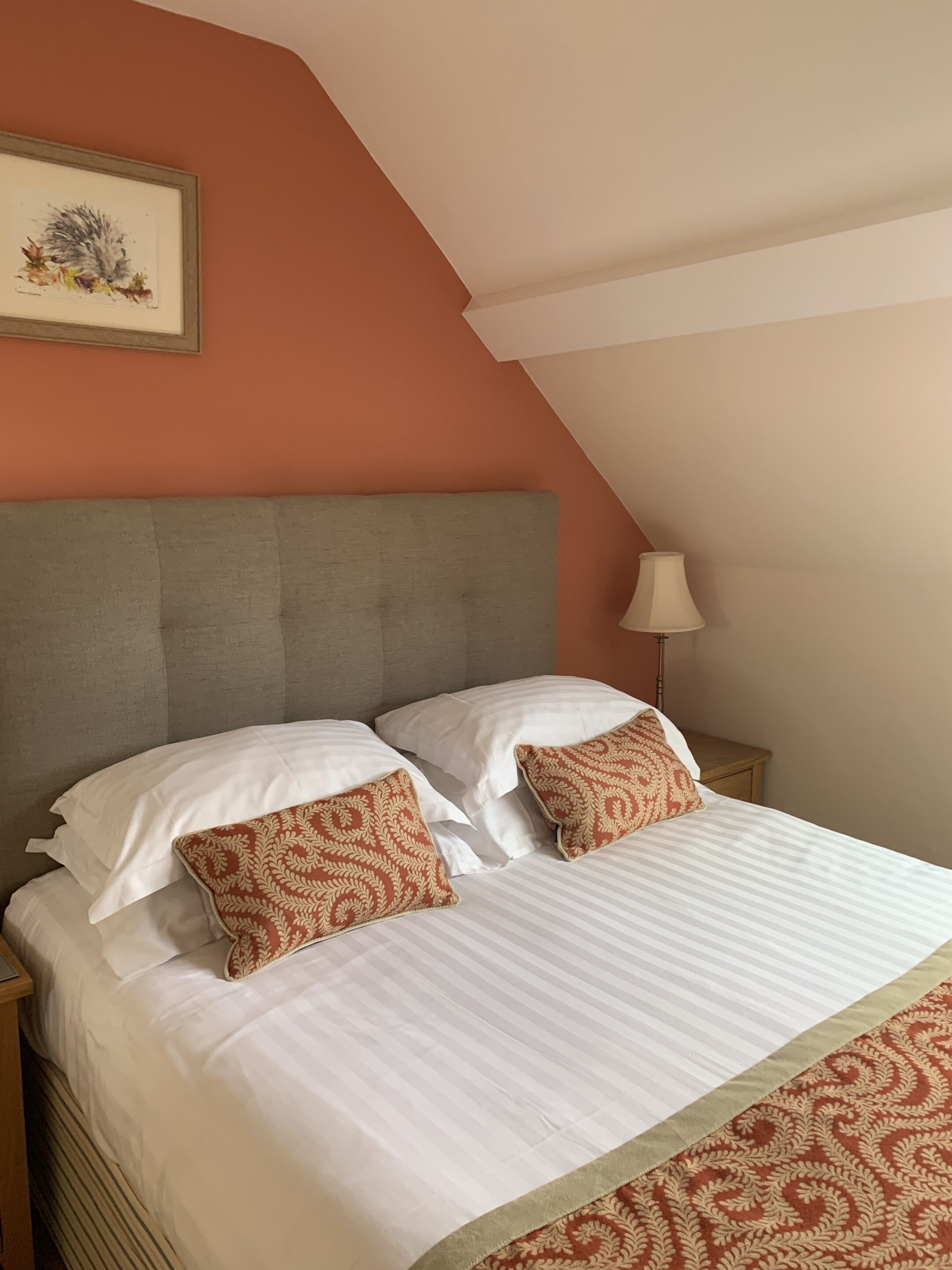 A neatly made bed with white linens and two patterned pillows, a grey upholstered headboard, and a framed artwork on the wall.