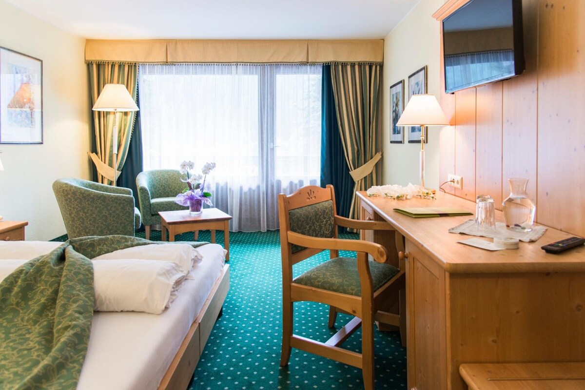 A hotel room with a bed, two armchairs, a desk, a TV, and a window with curtains.