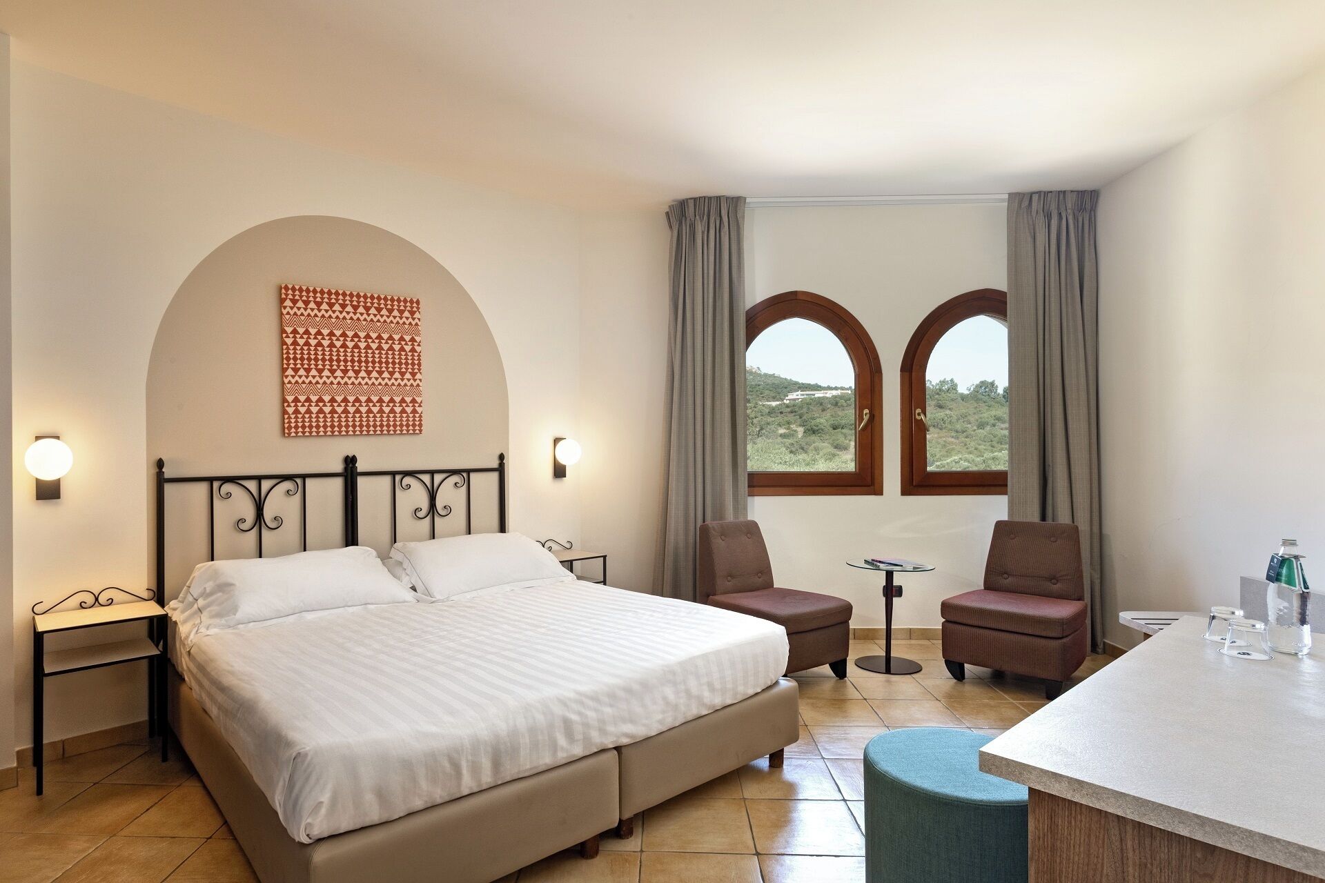 Habitación de hotel con cama, dos sillas, una mesita, un escritorio y vistas al exterior a través de dos ventanas.