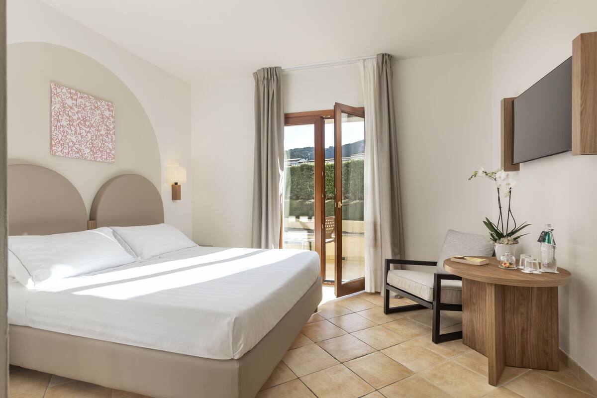 Ein Hotelzimmer mit Bett, Stuhl, kleinem Tisch mit Pflanze und einem an der Wand befestigten Fernseher.