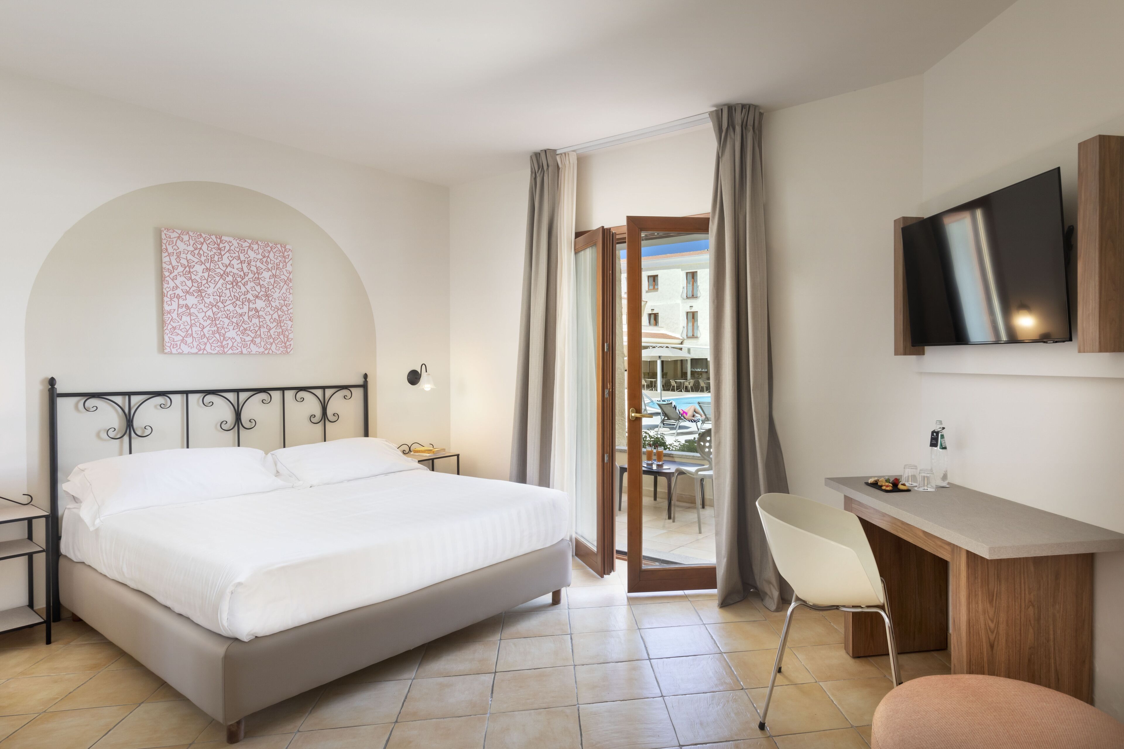 A hotel room with a bed, a desk, a chair, a TV, and a balcony with a view of buildings.