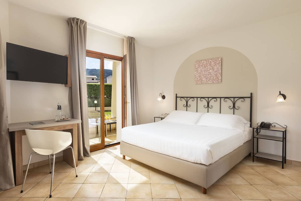 Ein Schlafzimmer mit Bett, Schreibtisch, Stuhl, Fernseher und einem Balkon mit Vorhängen.