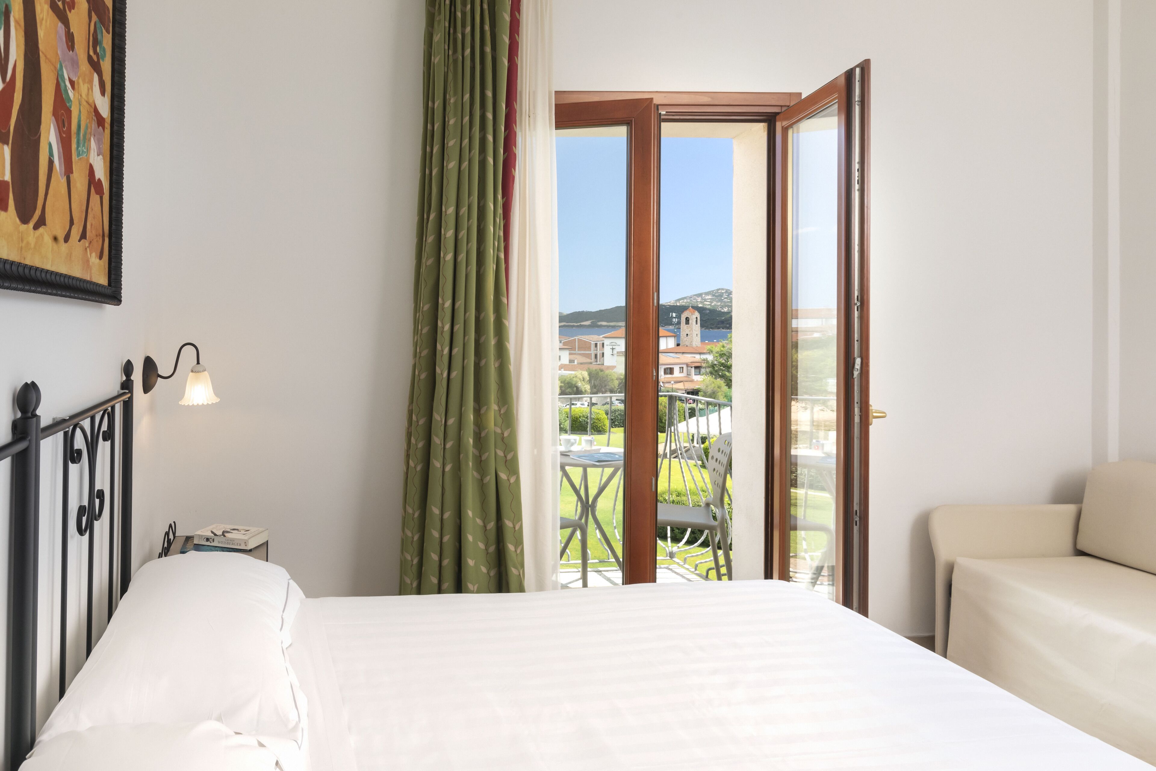 A bedroom with a bed, a nightstand, a lamp, a green curtain, and a view of a balcony with a table and chairs.