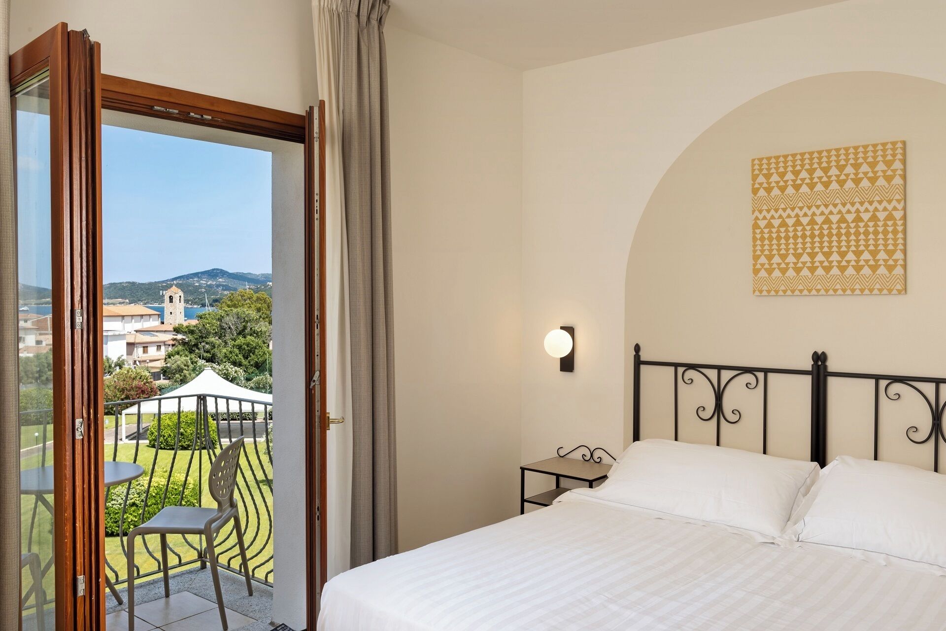 A hotel room with a bed, a nightstand, a wall-mounted light, a view of the outside, and a patterned artwork.