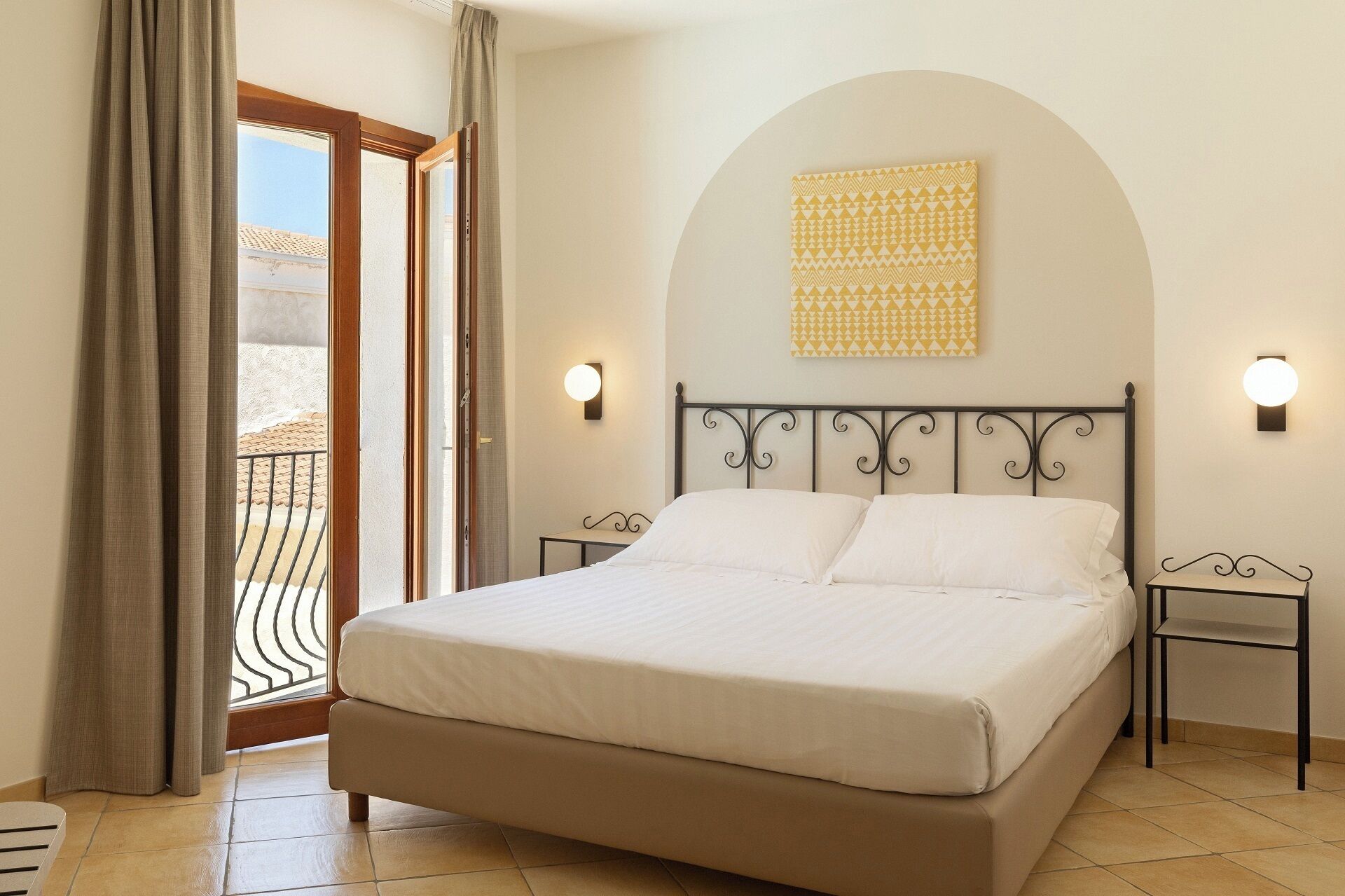 A bedroom with a bed, two bedside tables, a wall-mounted light fixture, and a view through a window with curtains.