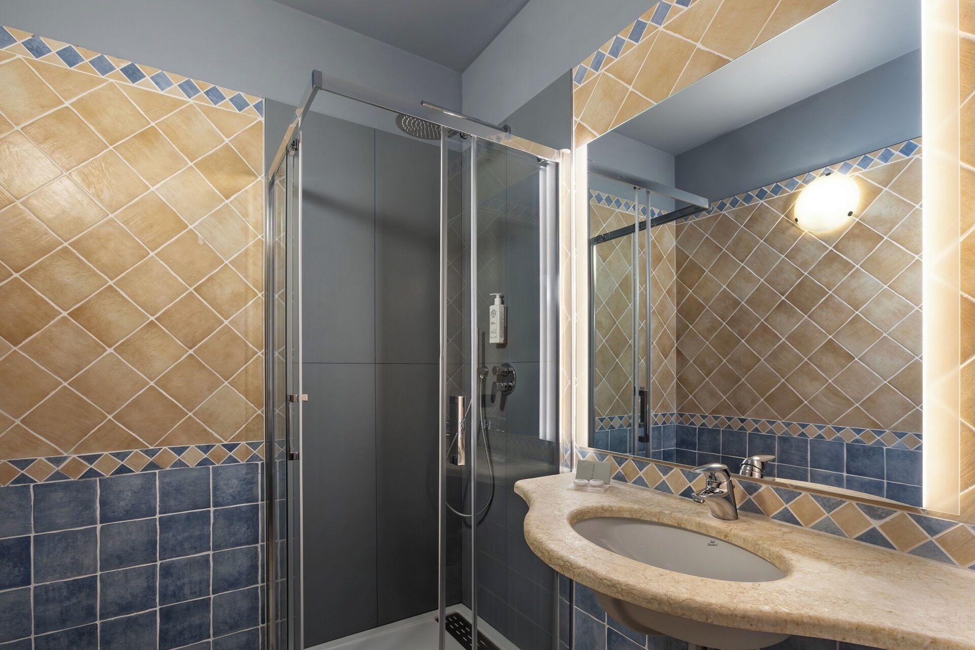 A bathroom with a glass-enclosed shower, a sink with a beige countertop, and tiled walls with a diamond pattern.