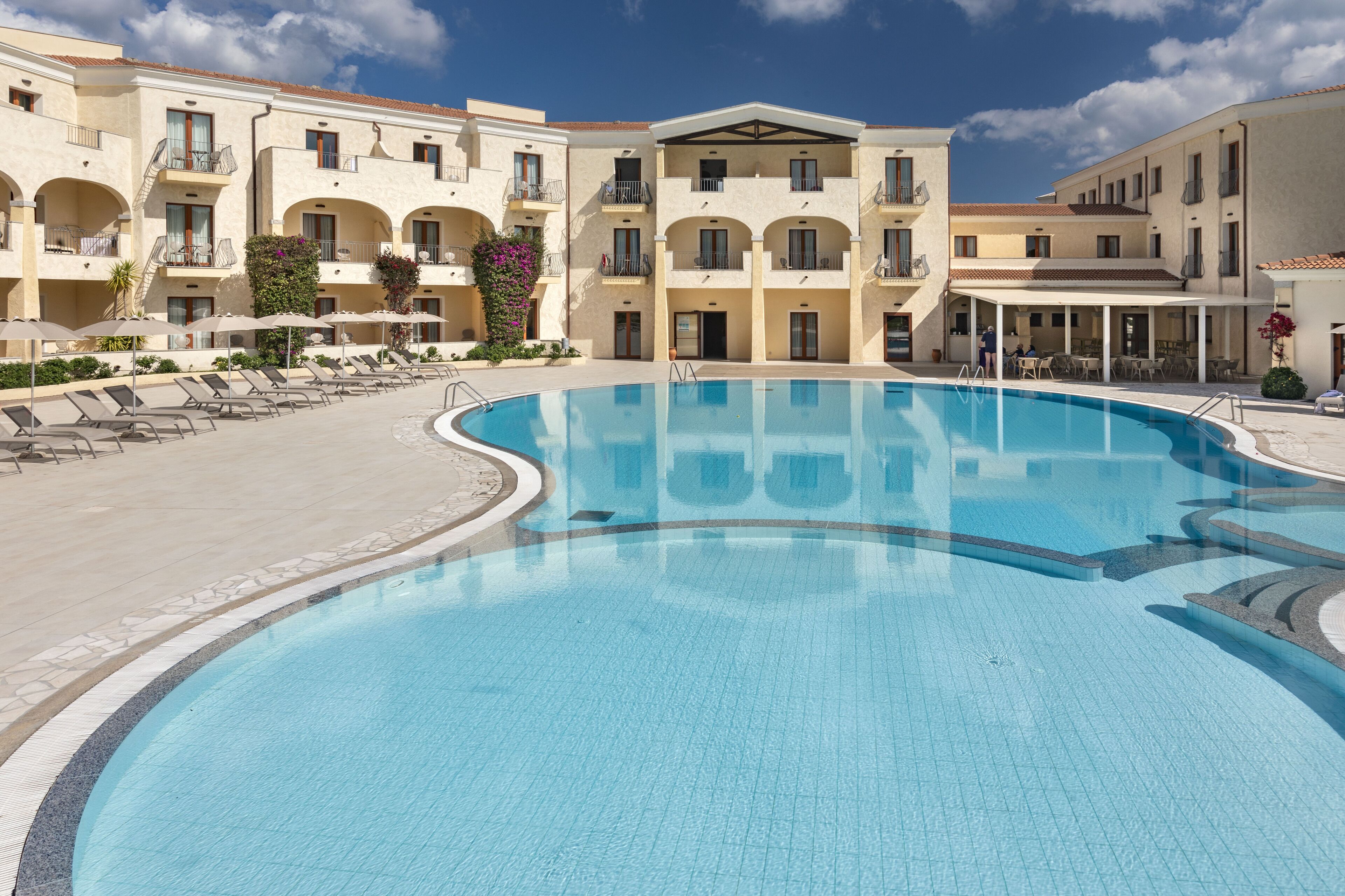 A swimming pool with sun loungers and a covered seating area, surrounded by a multi-story building with balconies and a tiled roof.