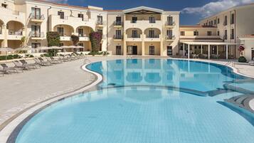 A swimming pool with sun loungers and a covered seating area, surrounded by a multi-story building with balconies and a tiled roof.