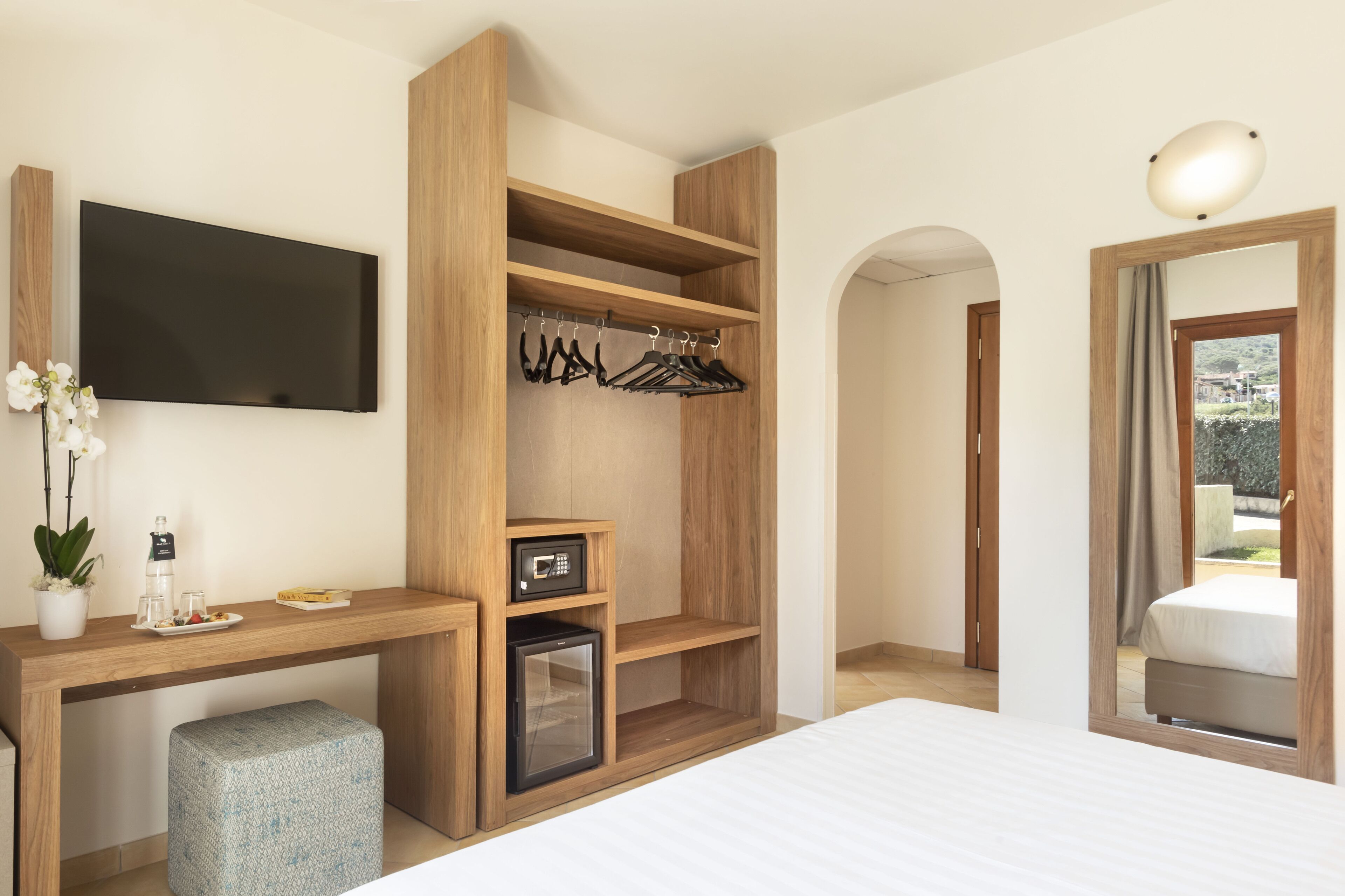 Une chambre d’hôtel moderne dotée d’un bureau en bois, d’un téléviseur à écran plat, d’un mini-réfrigérateur, d’un petit réfrigérateur, d’un lit et d’un dressing où sont suspendus des vêtements.
