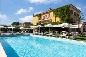 A swimming pool with lounge chairs and umbrellas, surrounded by a building with balconies and a clear sky.