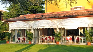 Restaurante com mesas ao ar livre, protegido por guarda-sóis brancos e cercado por vasos de plantas.