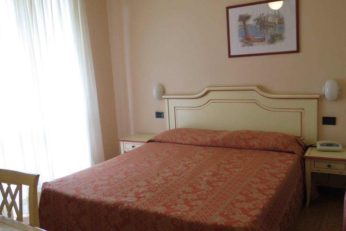 A hotel room with a bed, a desk, a chair, a nightstand, a bedside table, a lamp, a ceiling fan, and a framed picture on the wall.