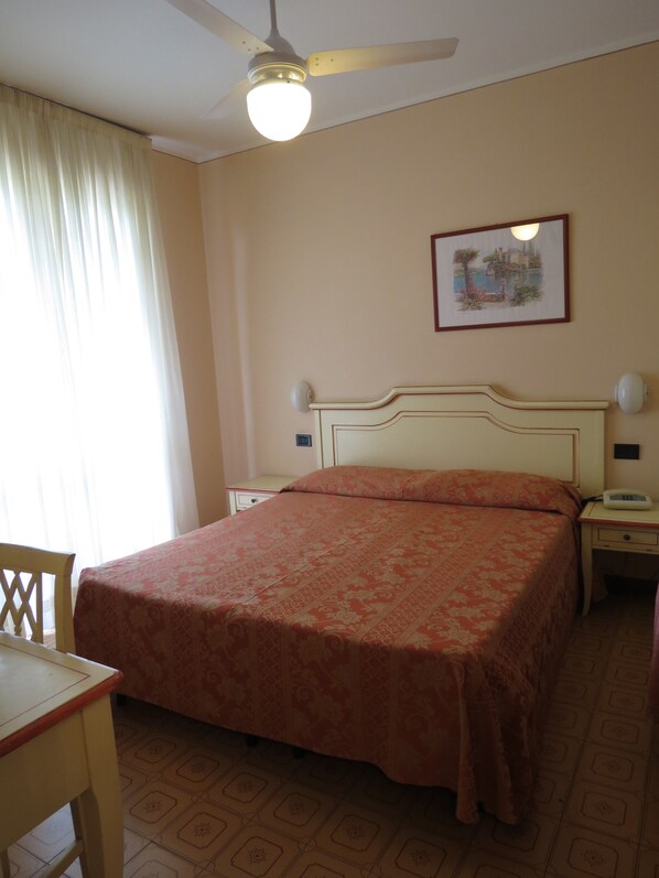 A hotel room with a bed, a desk, a chair, a nightstand, a bedside table, a lamp, a ceiling fan, and a framed picture on the wall. - La Quiete Park Hotel (Manerba del Garda)