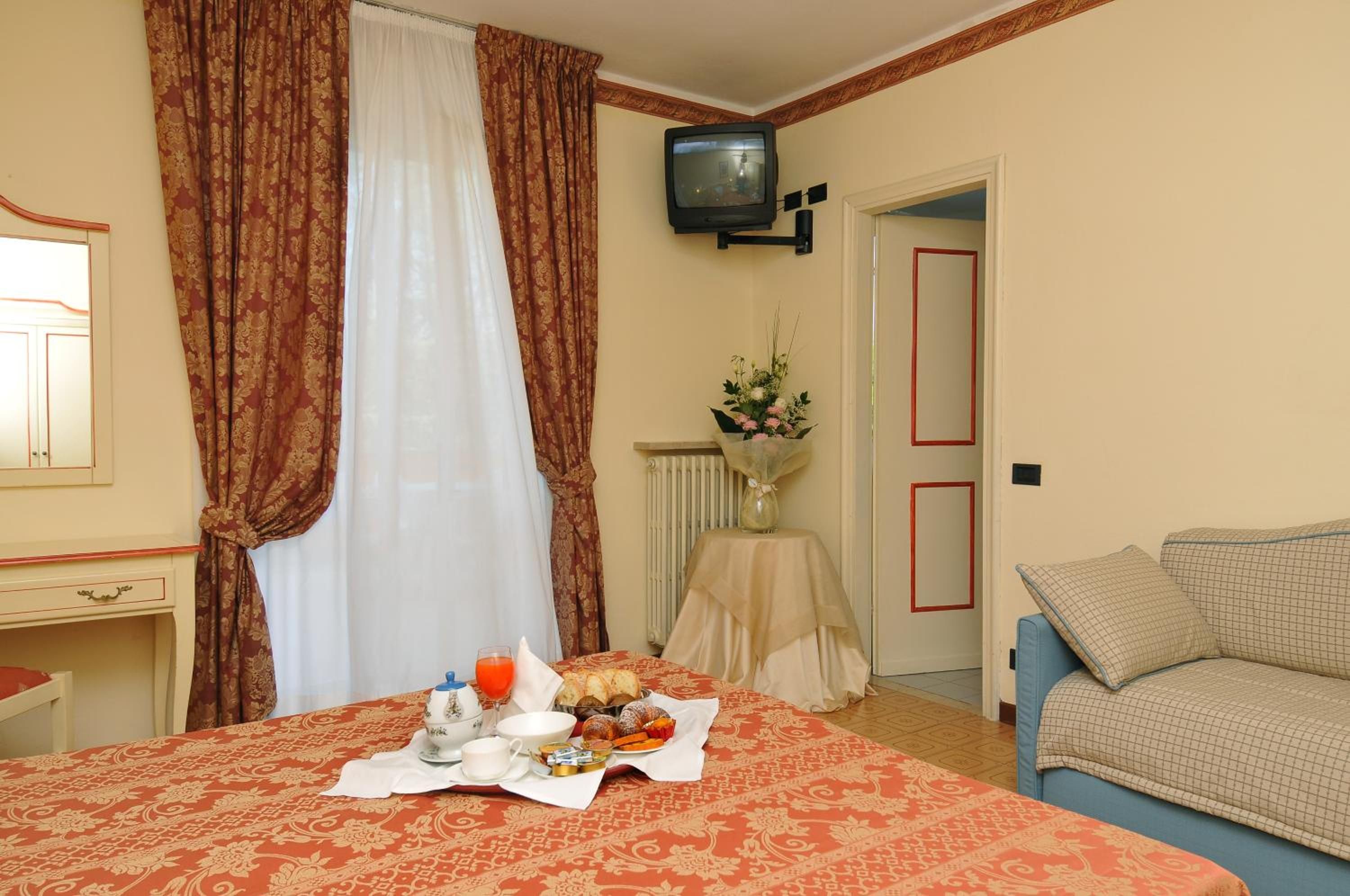 A hotel room with a bed, a television, a dining table with breakfast, a sofa, and a radiator.