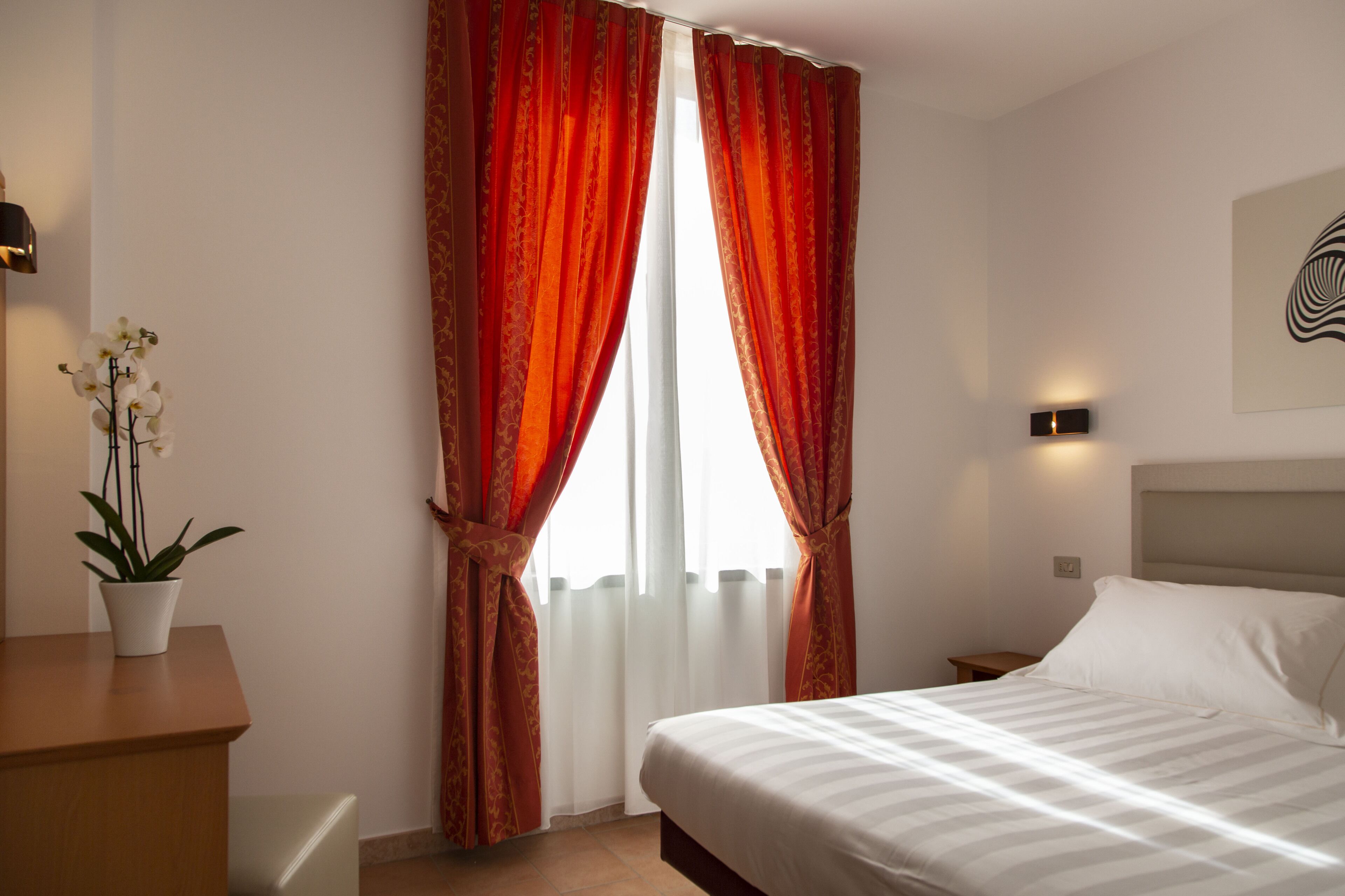 Una camera d'albergo con un letto, un comodino, una lampada, una pianta in vaso e una finestra con tende.
