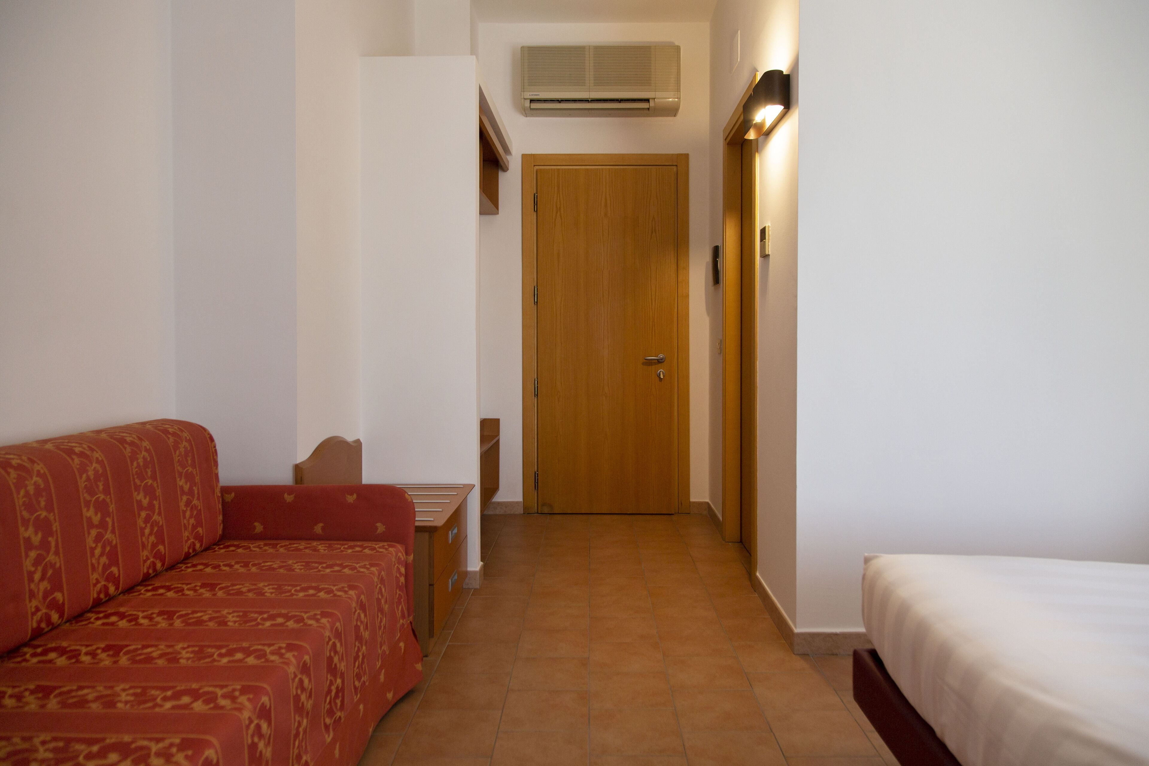 A hotel room with a wooden door, a red patterned sofa, a white bed, and a small table with a chair.
