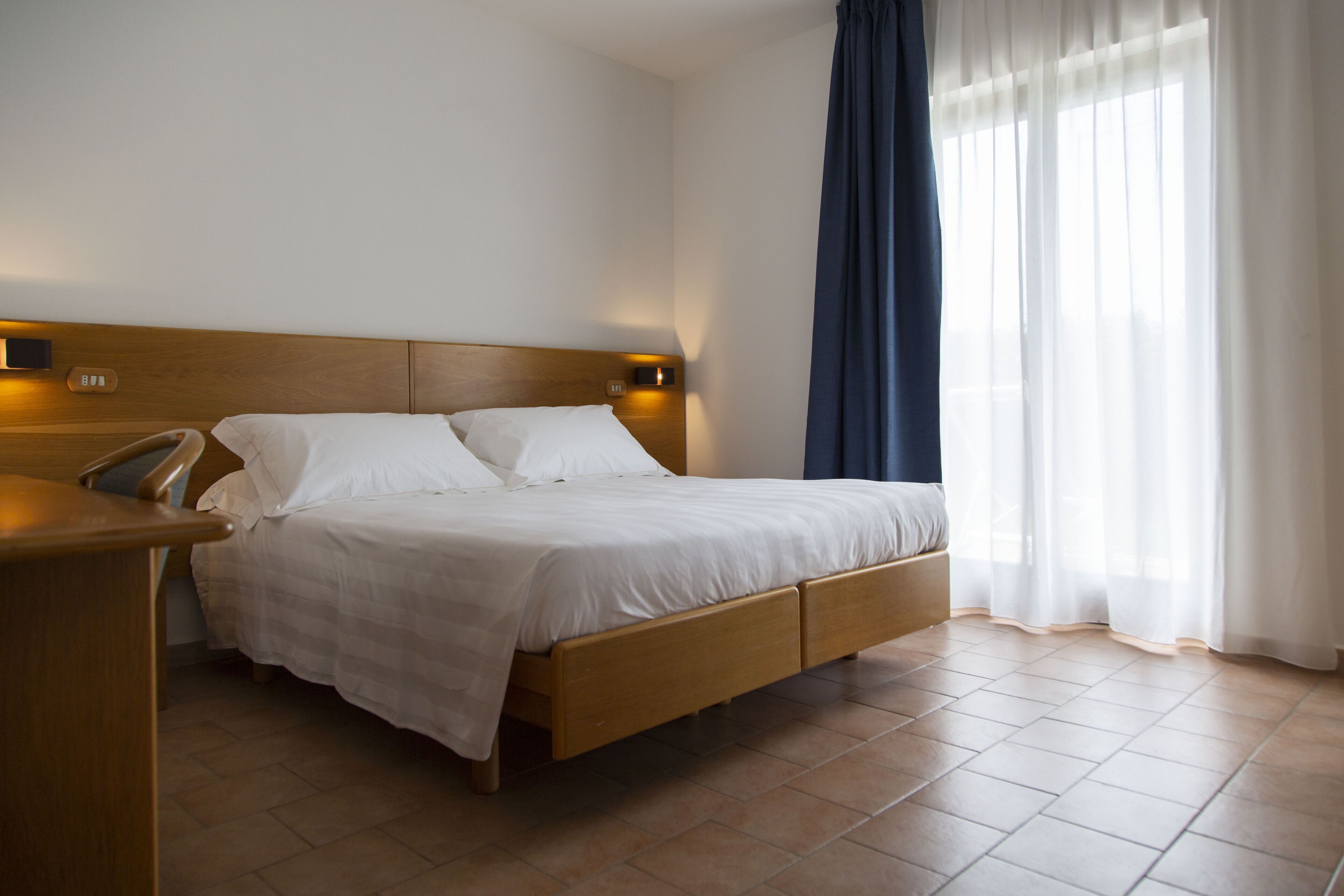 A modern hotel room with a wooden bed, a bedside table, and a large window with sheer curtains.