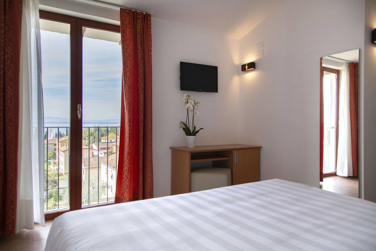 Ein Hotelzimmer mit einem Bett, einem Schreibtisch mit Topfpflanze, einem Fernseher und einem Balkon mit roten Vorhängen.