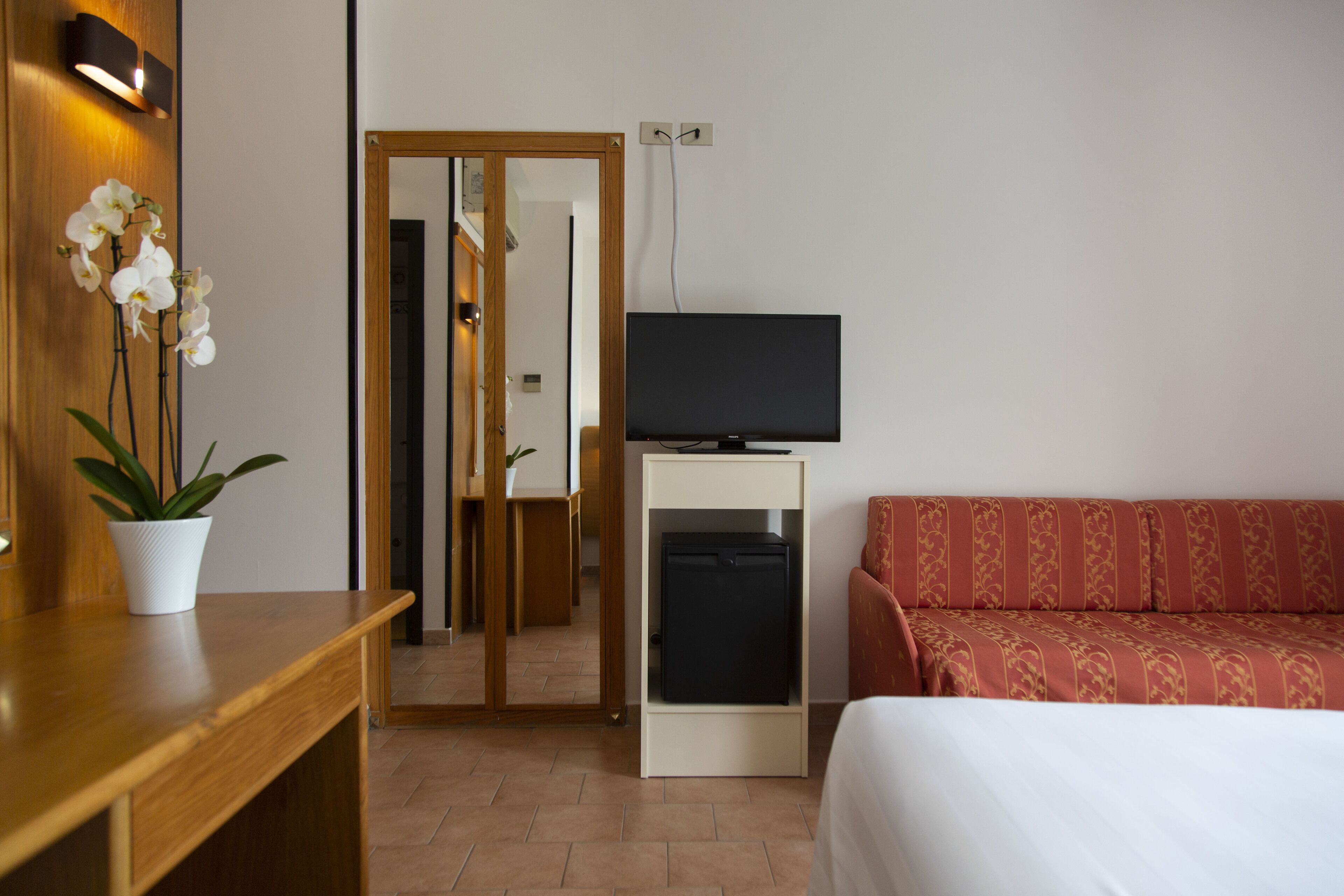 Habitación de hotel con escritorio de madera, planta en maceta, televisor sobre un mueble, cama con colcha estampada y sofá rojo.