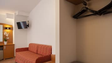 A compact room with a sofa, a small desk, a flat-screen TV mounted on the wall, and a compact shelving unit.