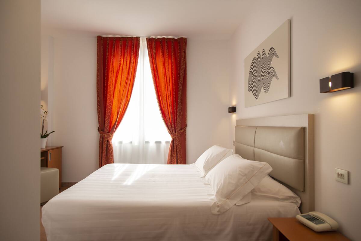 Ein Hotelzimmer mit einem großen Bett, einem Nachttisch mit Lampe, einem Wandbild und einem Fenster mit roten Vorhängen.