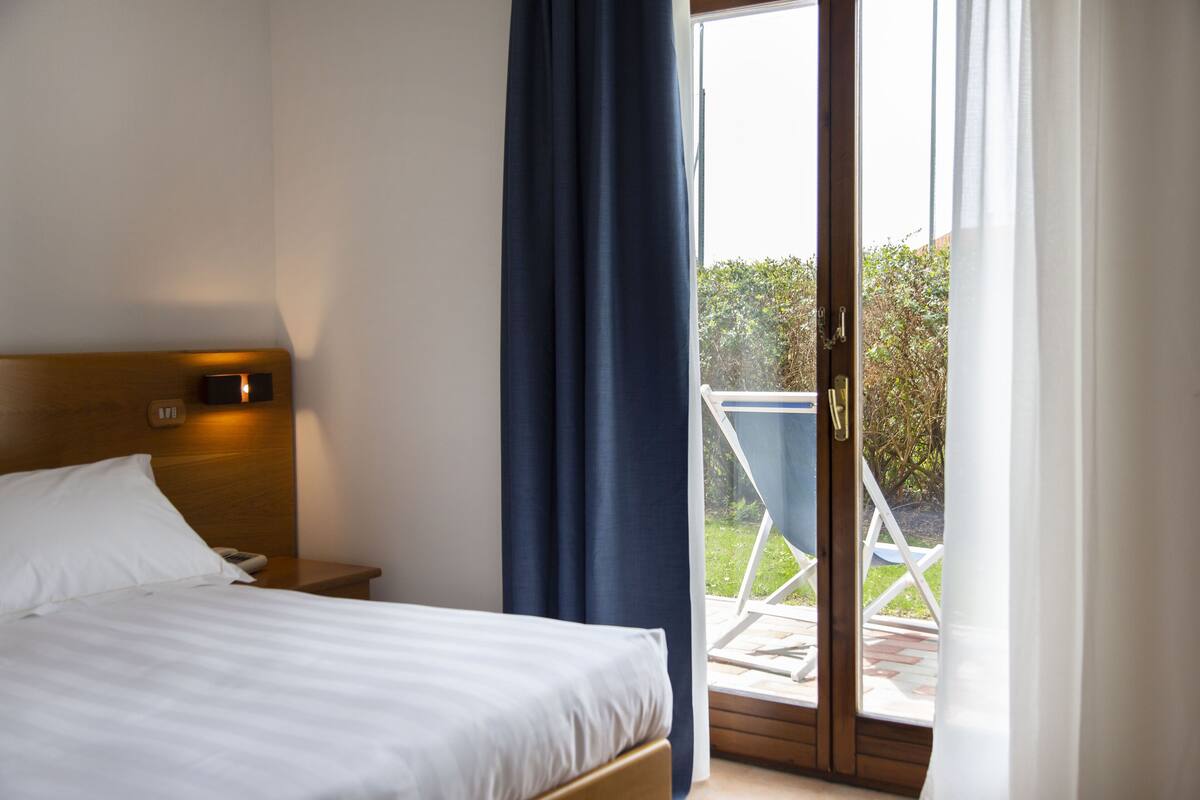 Ein Hotelzimmer mit einem Bett, einem Nachttisch, einer Wandleuchte und Blick ins Freie durch eine Glastür.