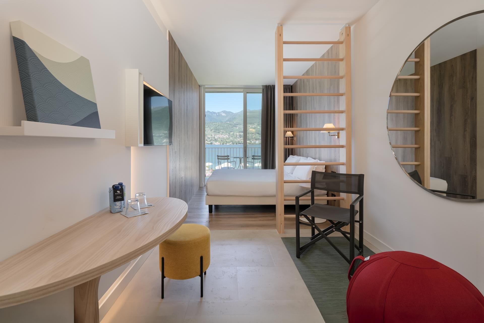 A modern hotel room with a bed, a desk, a chair, a small stool, a mirror, and a view of the mountains.