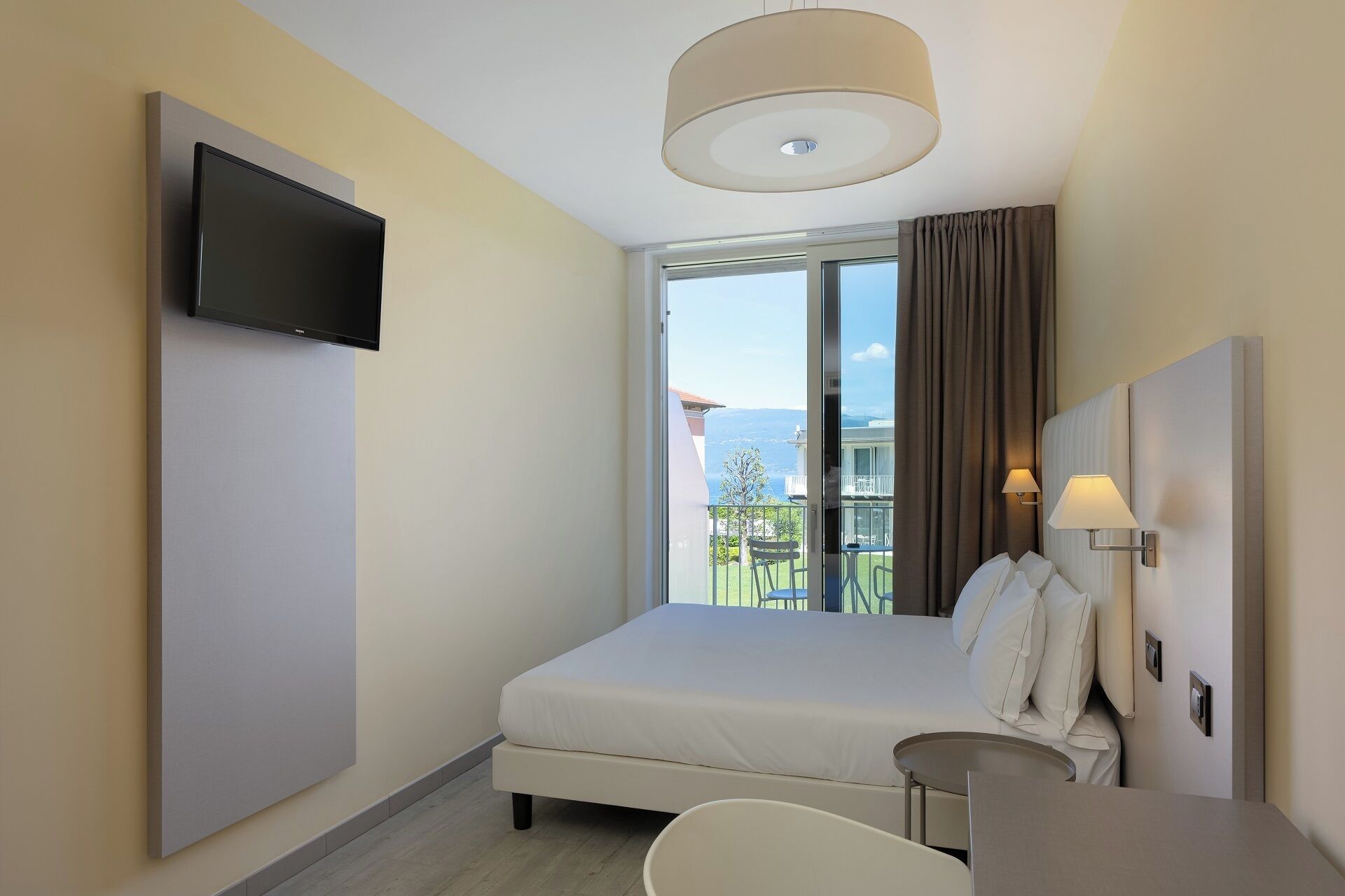 A hotel room with a bed, a TV mounted on the wall, a balcony with a view, and a table with a chair.