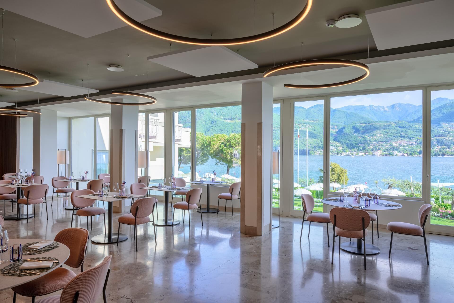 A restaurant with round tables and chairs, large windows, and a view of a lake and mountains.