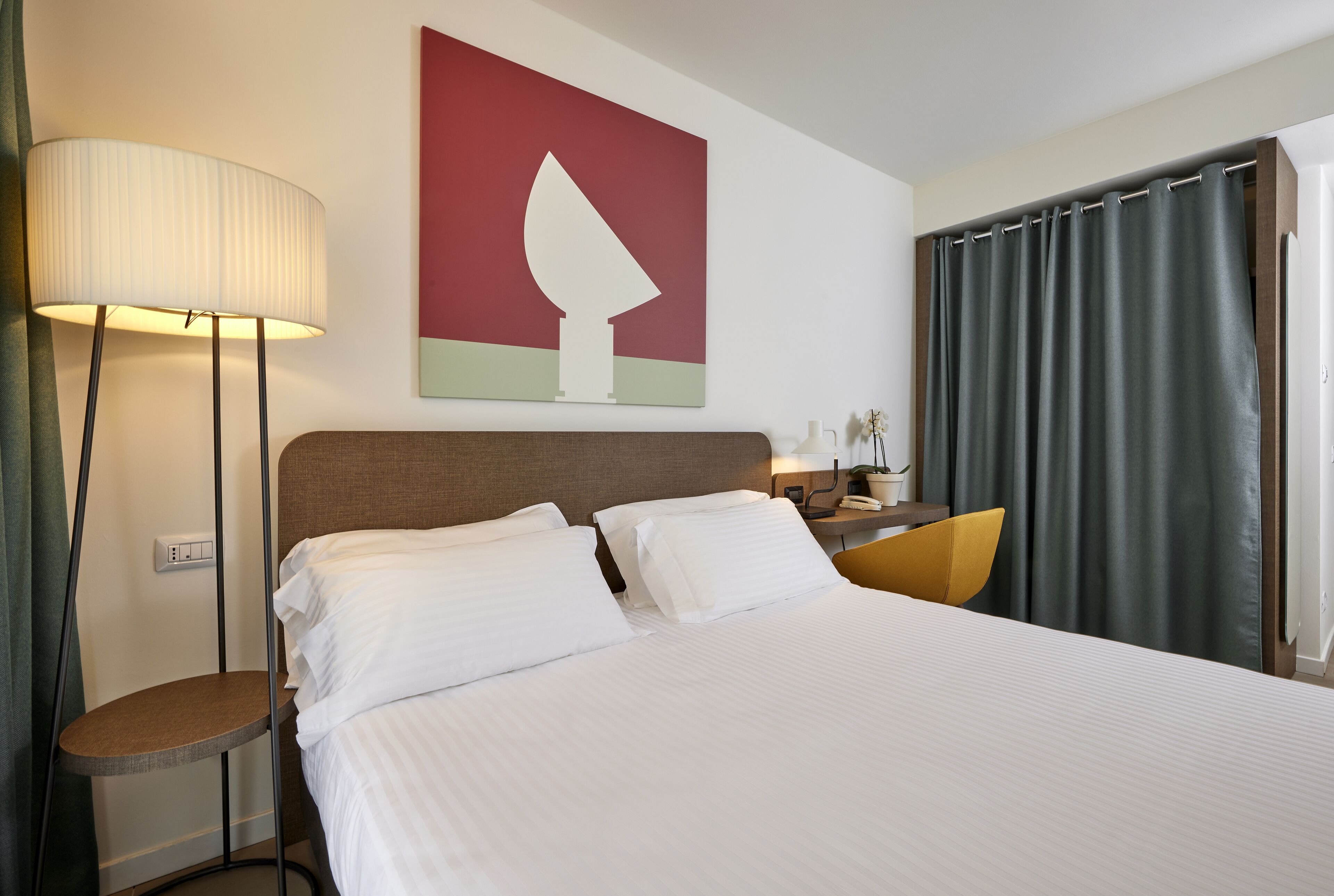 A hotel room with a large bed, a bedside table, a lamp, a chair, and a painting on the wall.