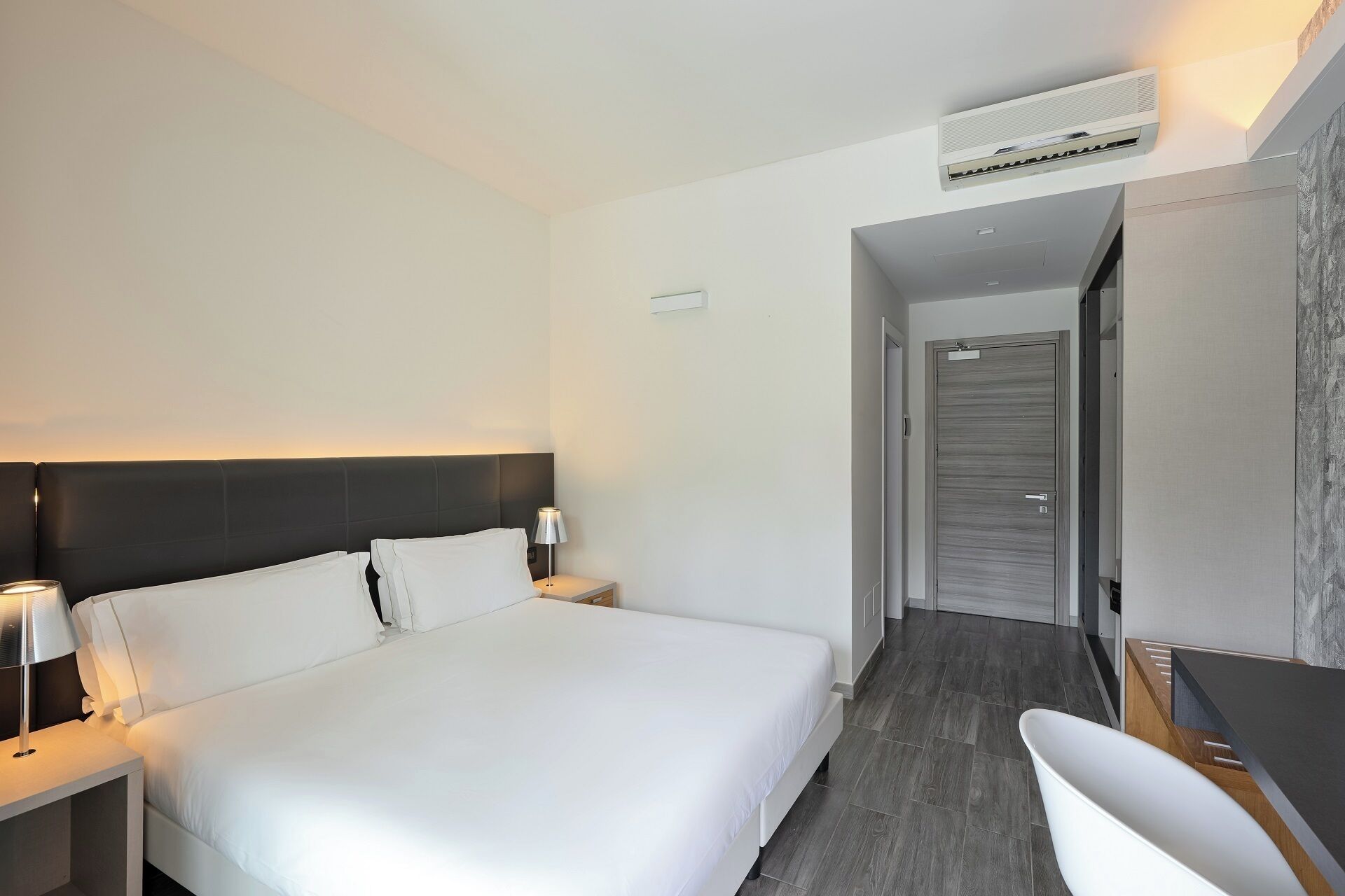 Habitación de hotel con una cama grande, mesitas de noche, un escritorio, una silla, un equipo de aire acondicionado y una puerta que da a otra habitación.