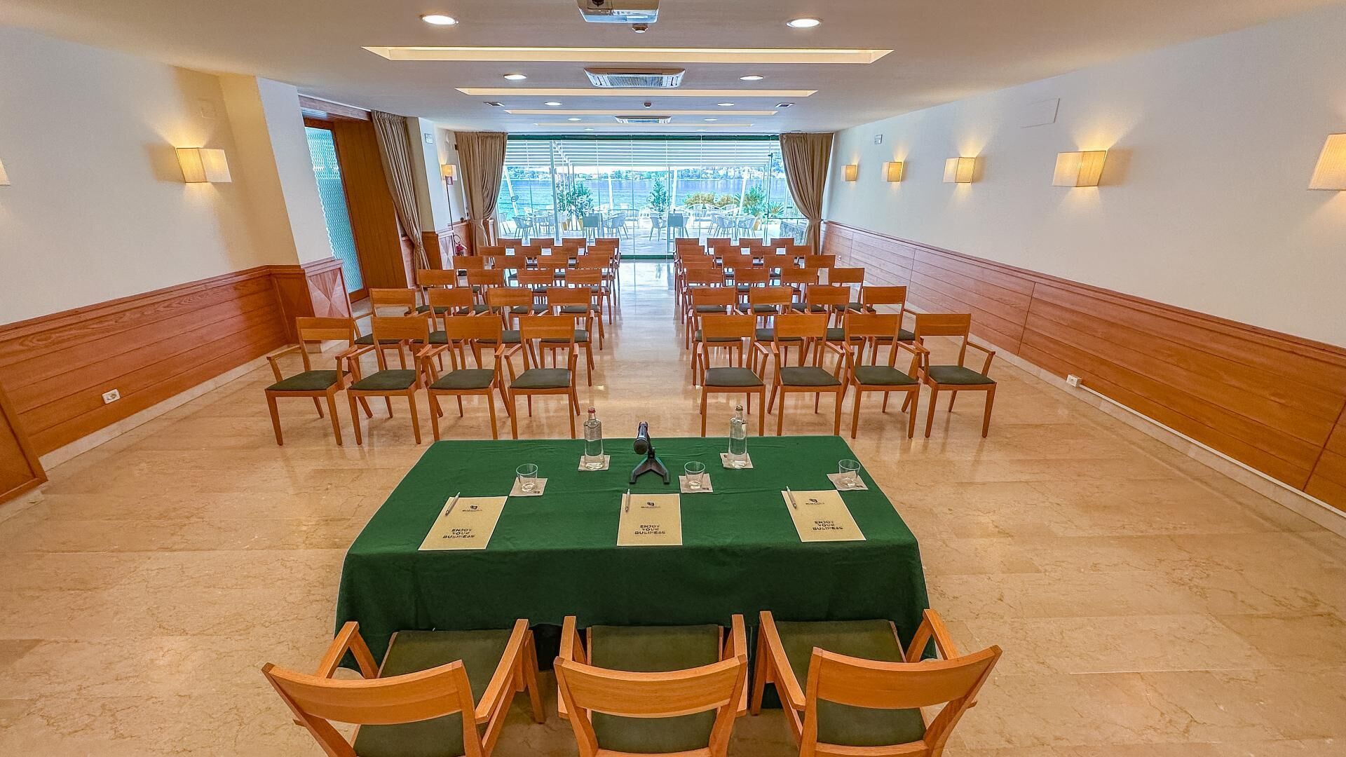Ein Konferenzraum mit Reihen aus Holzwstühlen, einem grünen Tisch mit Namensschildern und Blick ins Freie durch große Fenster.