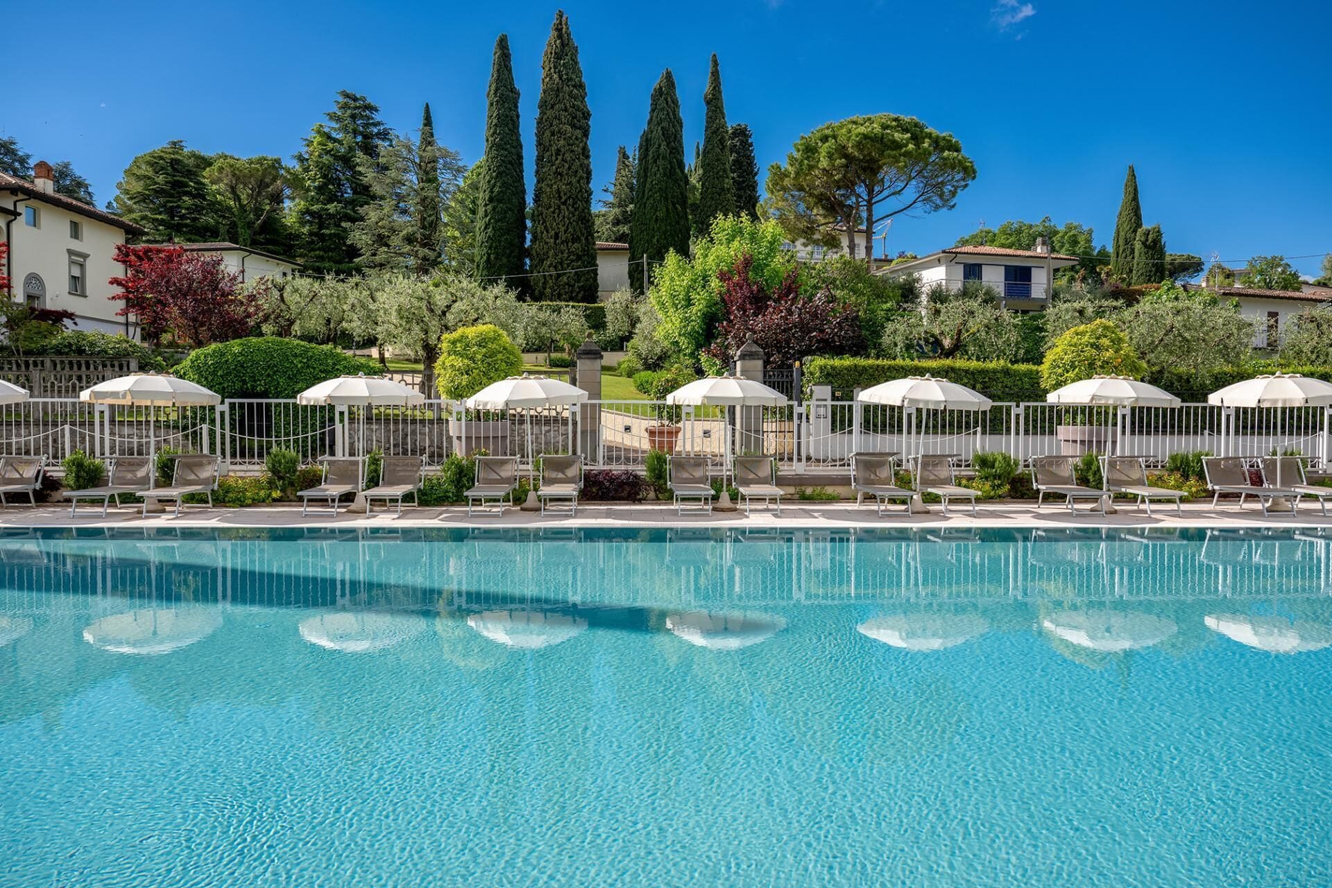 A swimming pool with lounge chairs and umbrellas, surrounded by greenery and tall trees.