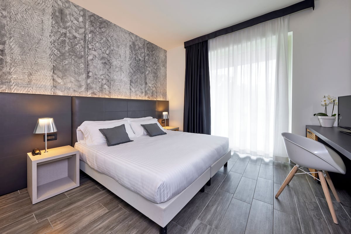 A modern hotel room with a large bed, bedside lamps, a desk, and a view of the city through sheer curtains.