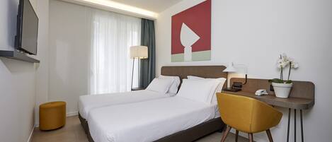 A hotel room with a bed, a desk with a chair, a TV, a lamp, and a plant.