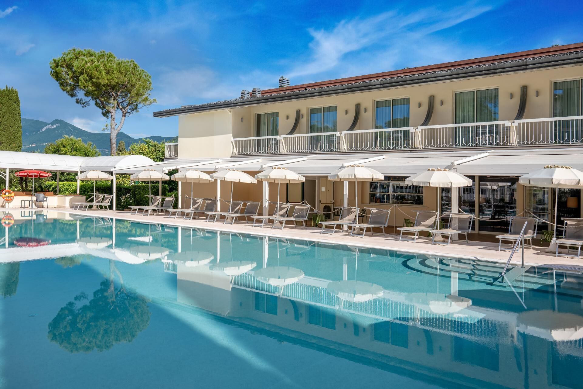 Une piscine avec des chaises longues et des parasols, un bâtiment doté d’un balcon, et une montagne en arrière-plan.