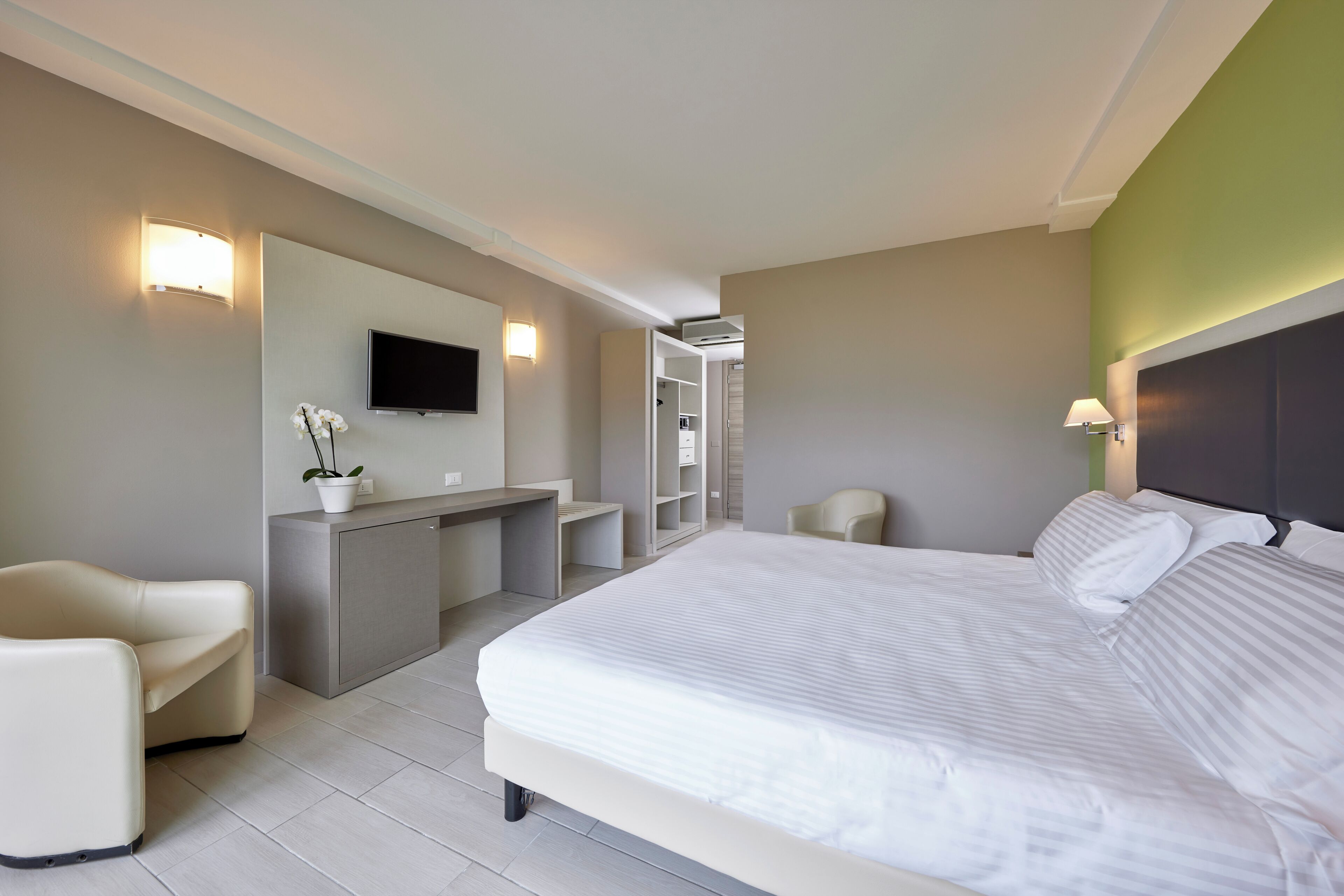 A modern hotel room with a large bed, a desk, a chair, a TV, and a wall-mounted lamp.