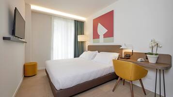 A modern hotel room with a large bed, a desk with a chair, a television, a lamp, and a potted plant.