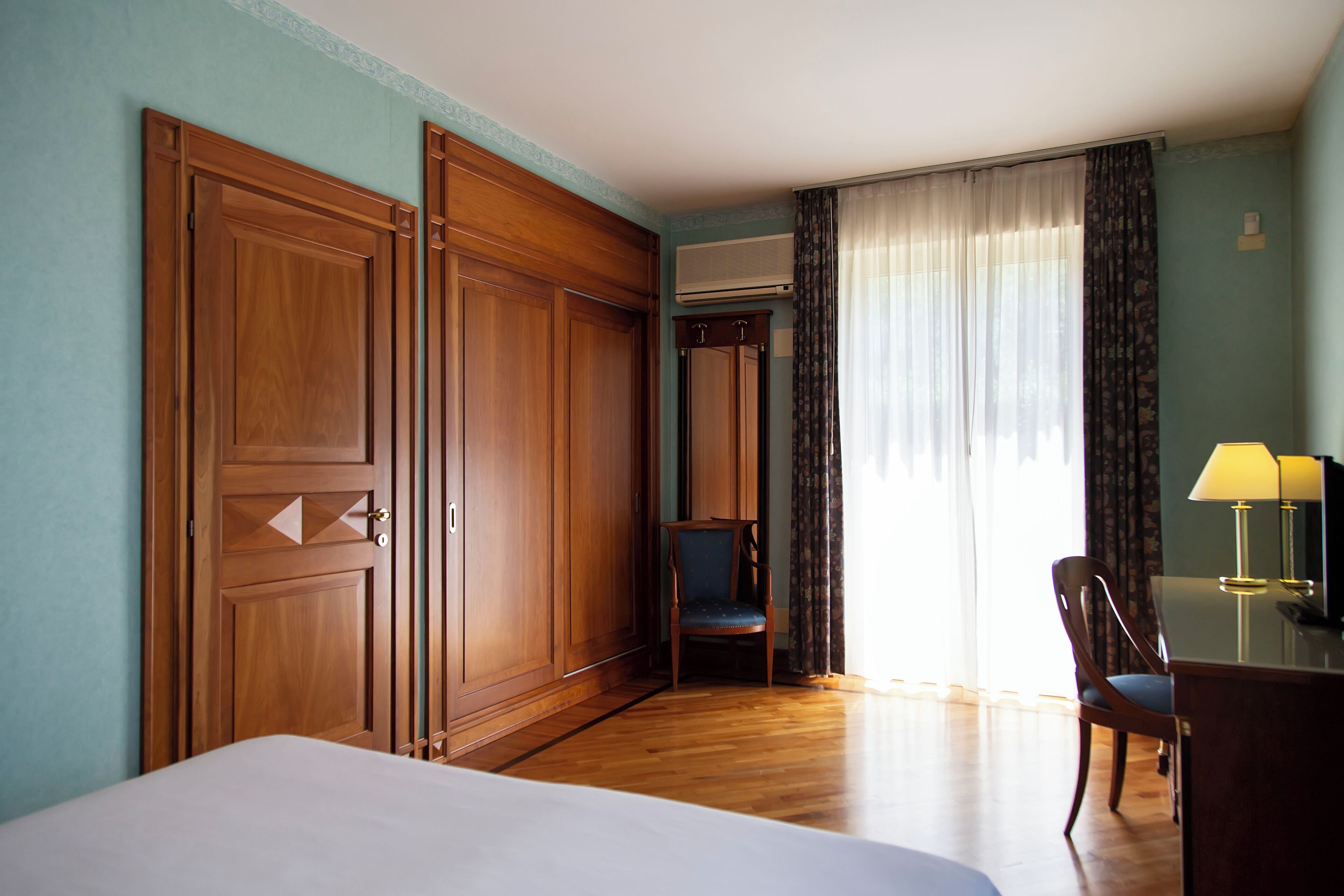 Ett hotellrum med en säng, en garderob i trä, ett skrivbord med en lampa, en stol och ett fönster med gardiner.