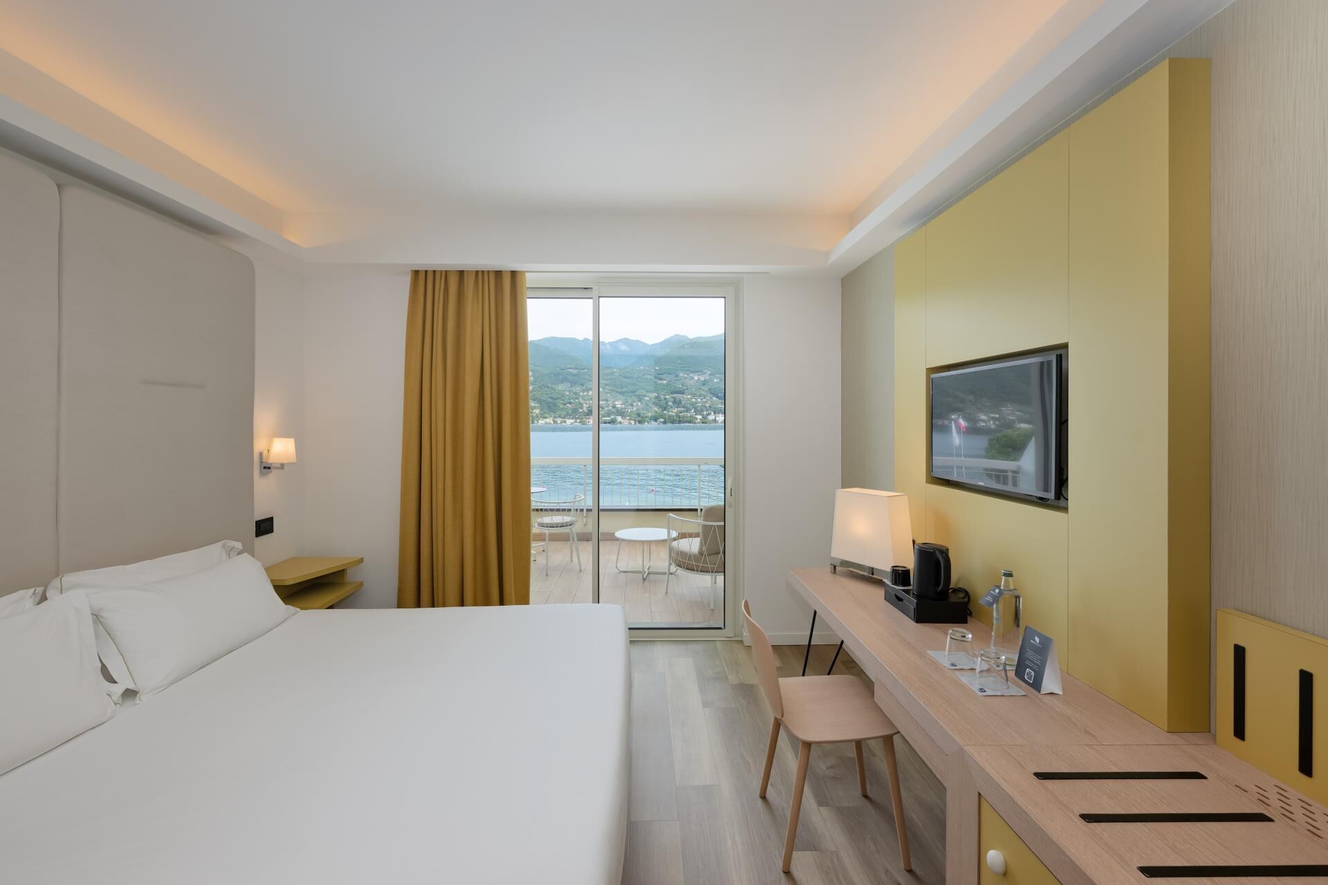 A hotel room with a bed, a desk with a chair, a television, and a view of a body of water and mountains.