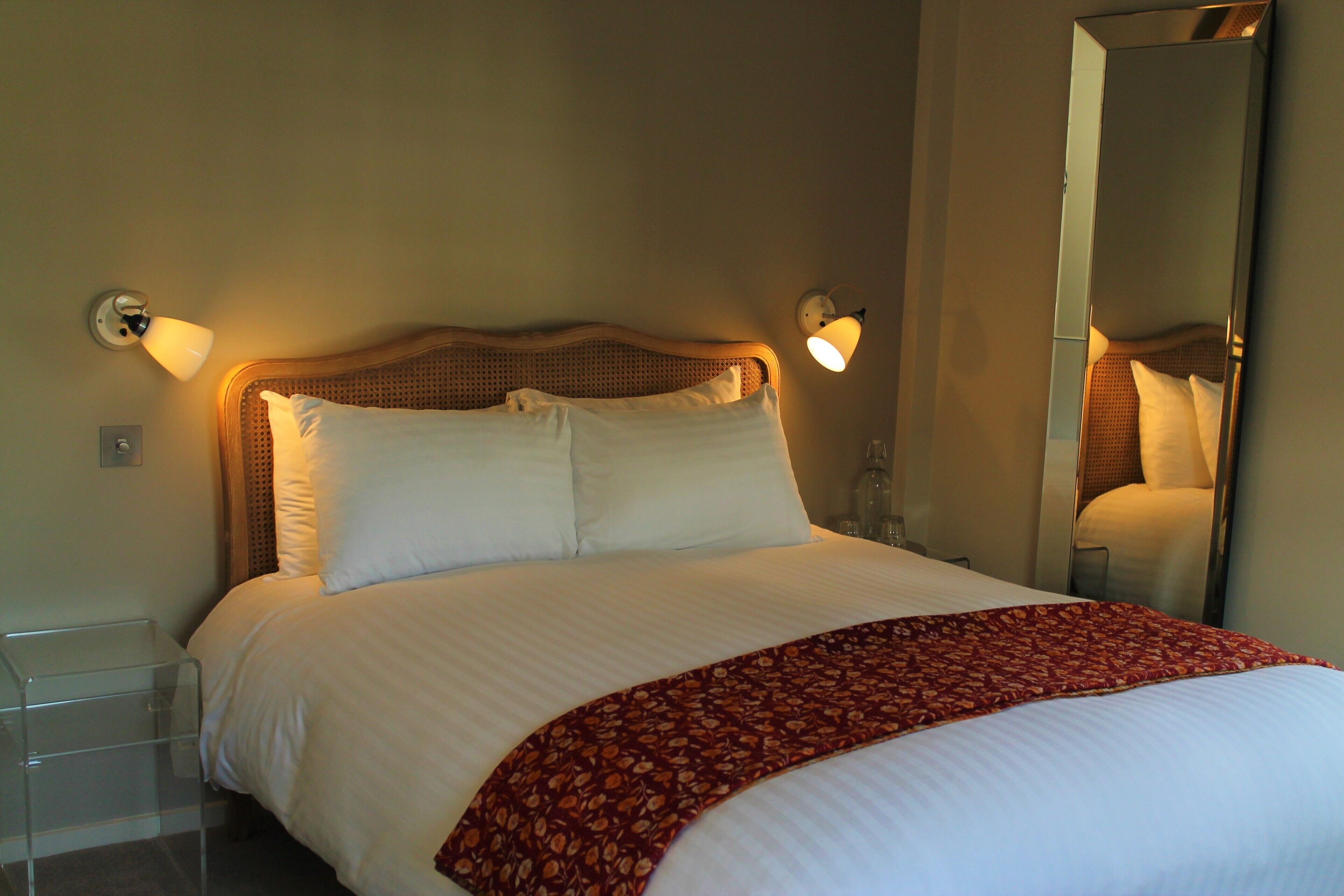 Een net opgemaakt bed met witte lakens en een rode woondeken, twee wandlampen voor het lezen en een grote spiegel aan de muur.