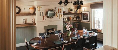 A dining room with a round table set for six, surrounded by chairs. There is a fireplace, a mirror, and a shelf with decorative items.