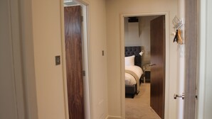 A narrow hallway leading to a bedroom with a tufted headboard bed, a wooden door on the left, and a coat rack with hooks on the right.