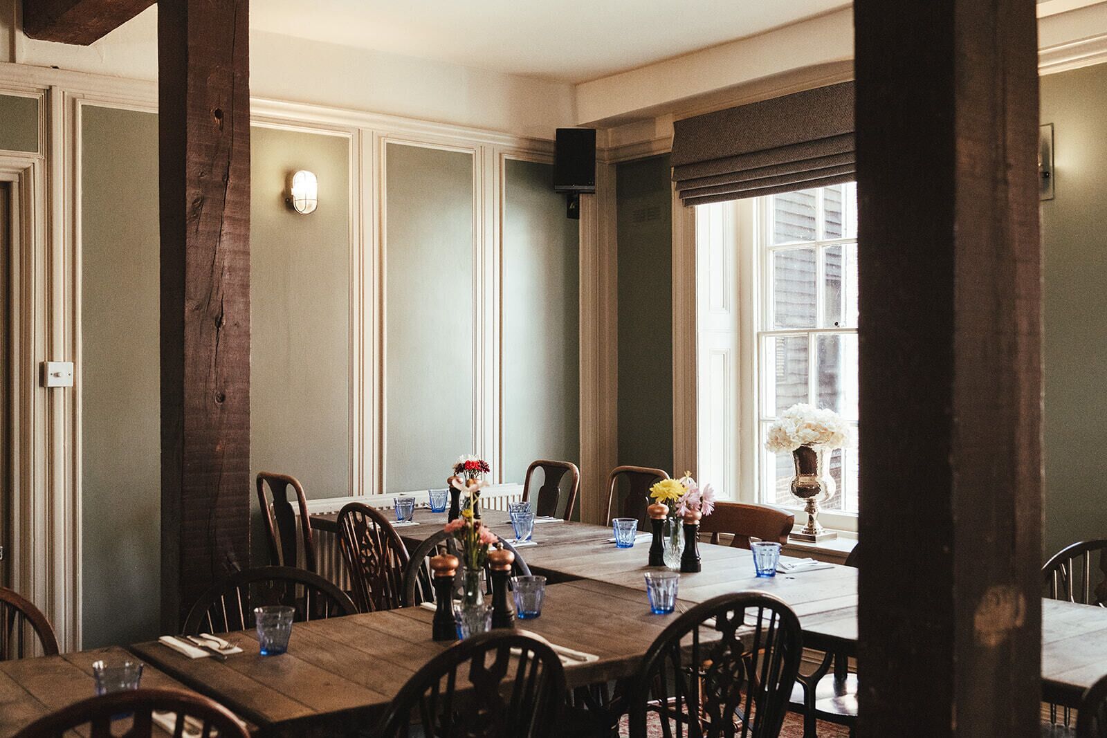 Uma sala de jantar com uma longa mesa de madeira posta para seis pessoas, rodeada de cadeiras. Há arranjos florais sobre a mesa e uma janela com persianas à direita.