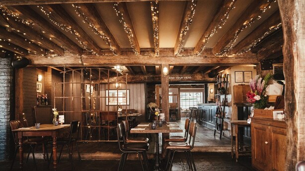 A rustic dining area with wooden tables and chairs, string lights on the ceiling, and a brick wall. - The Ship Inn (Rye)