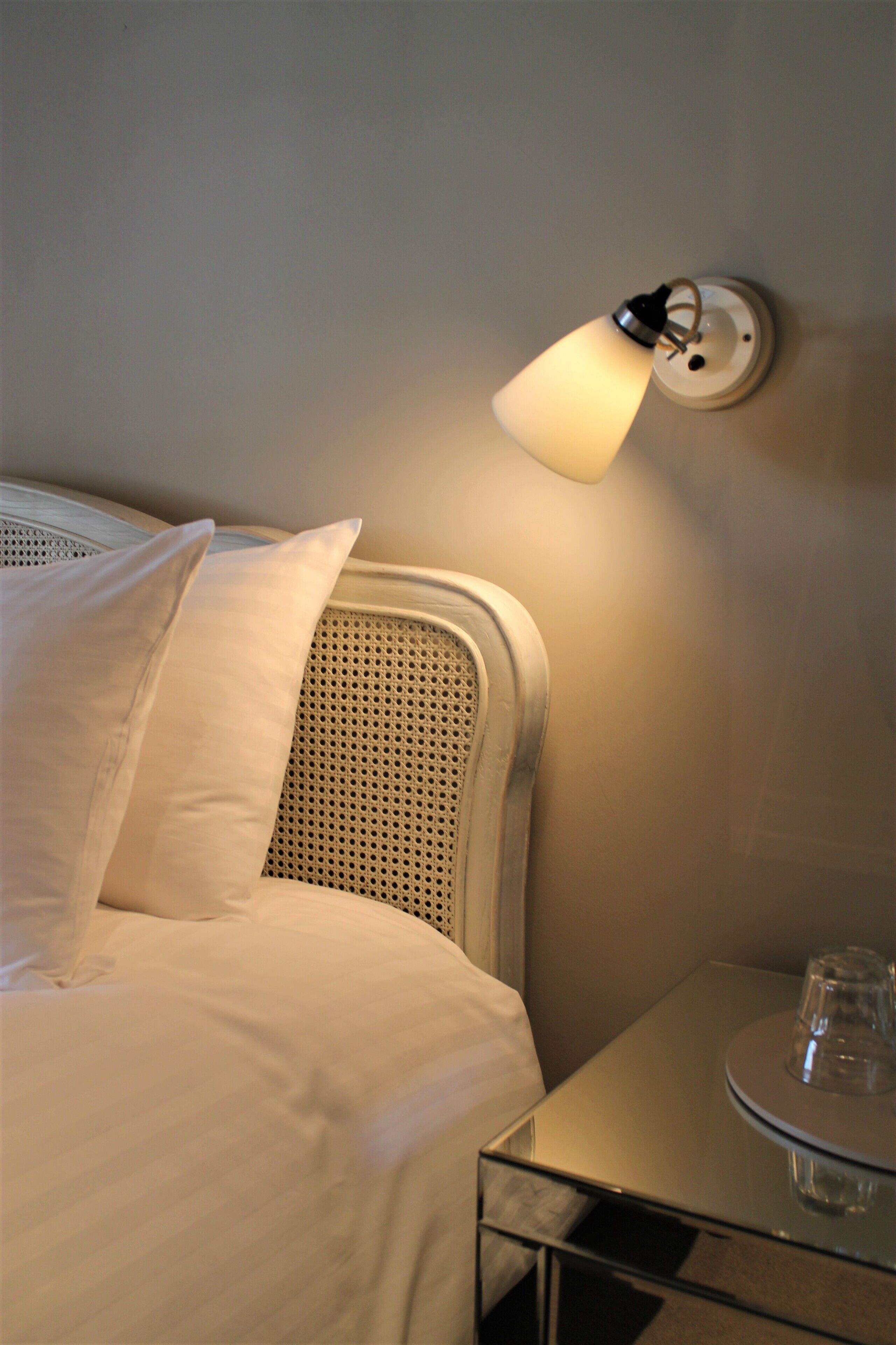 A neatly made bed with white linens and pillows, a wall-mounted lamp, and a small table with a glass on it.