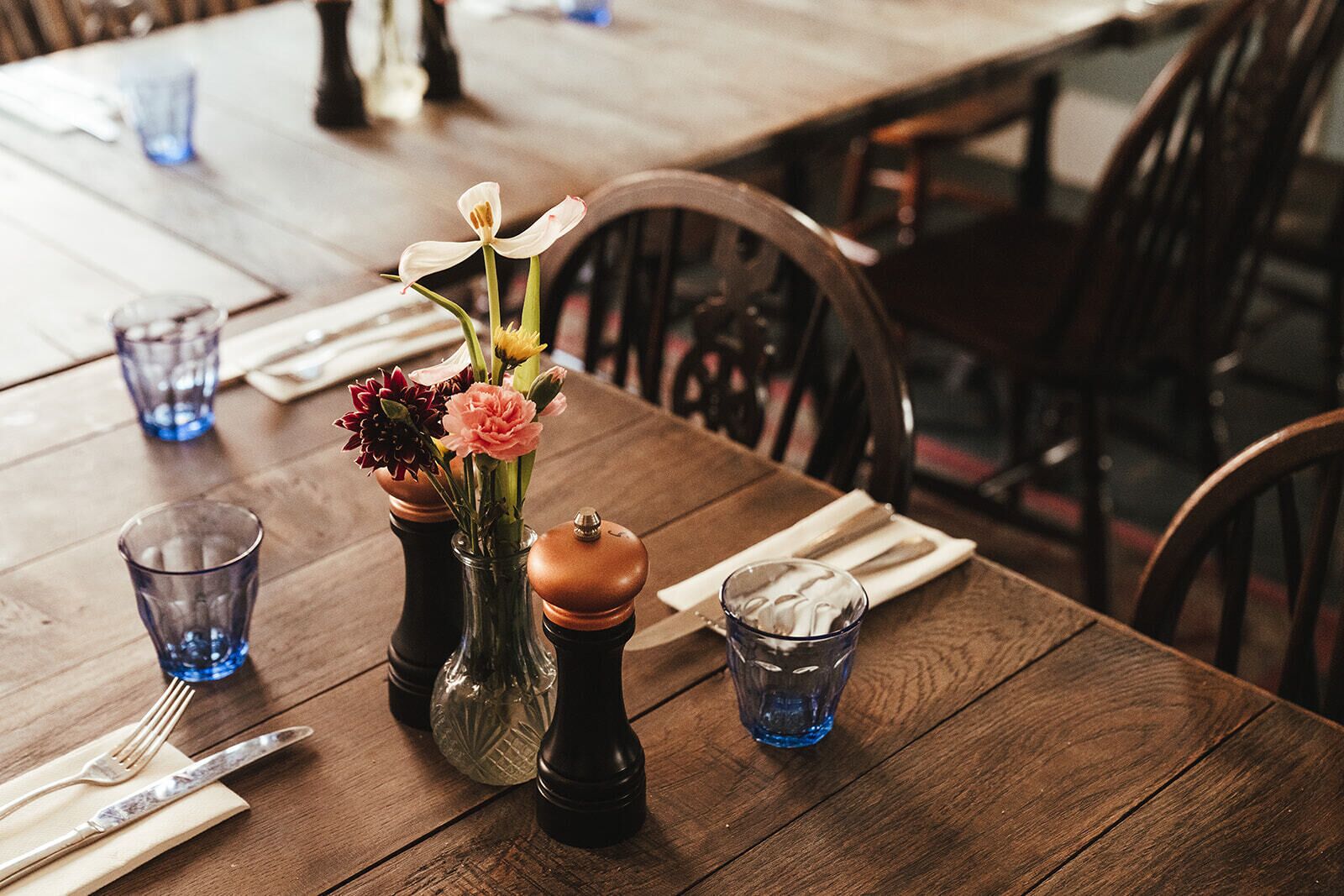 Una mesa de madera para comer con un florero de flores, saleros y pimenteros, y vasos de vidrio azules.