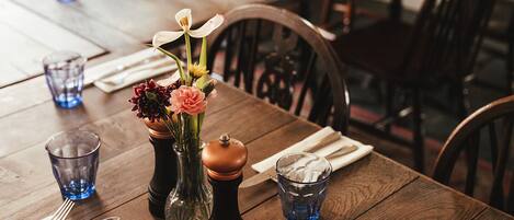Una mesa de madera para comer con un florero de flores, saleros y pimenteros, y vasos de vidrio azules.